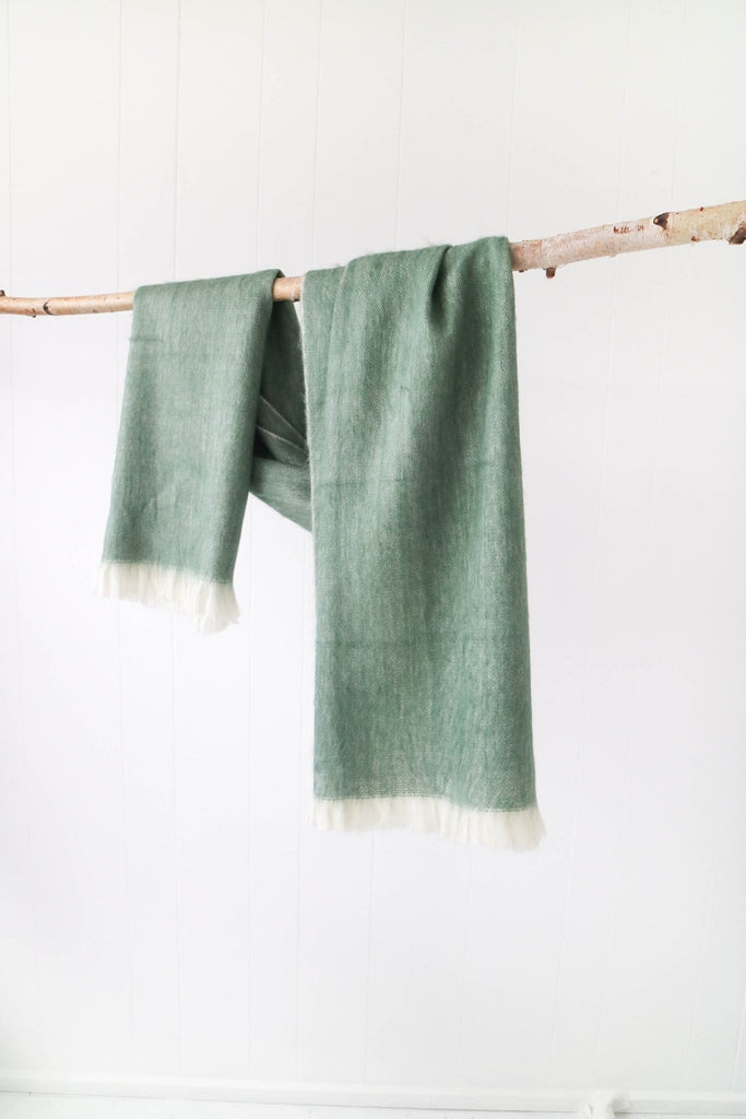 A sage alpaca scarf draped over a wooden branch in front of a white background