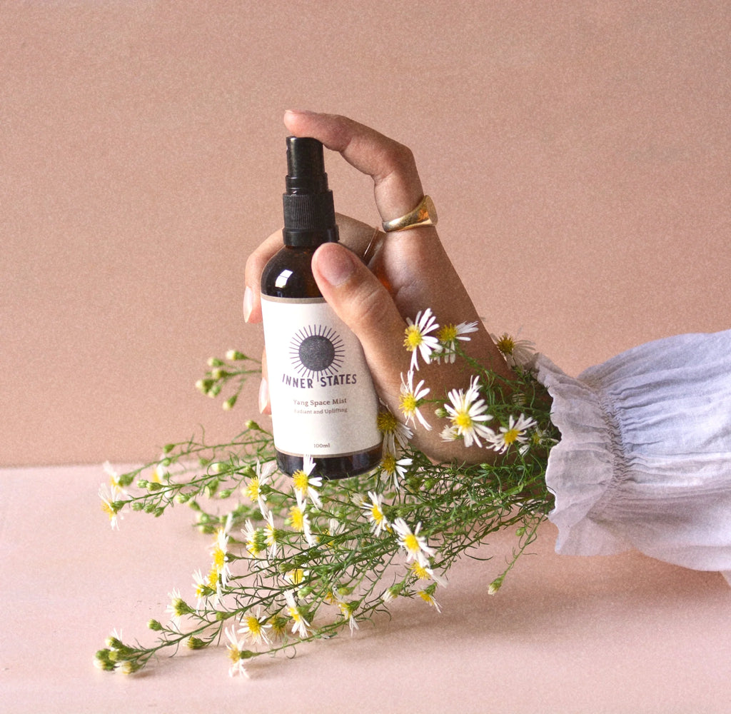 a person's hand holding a brown yang mist bottle by the brand Inner States. There's white daises around the product and a peach background